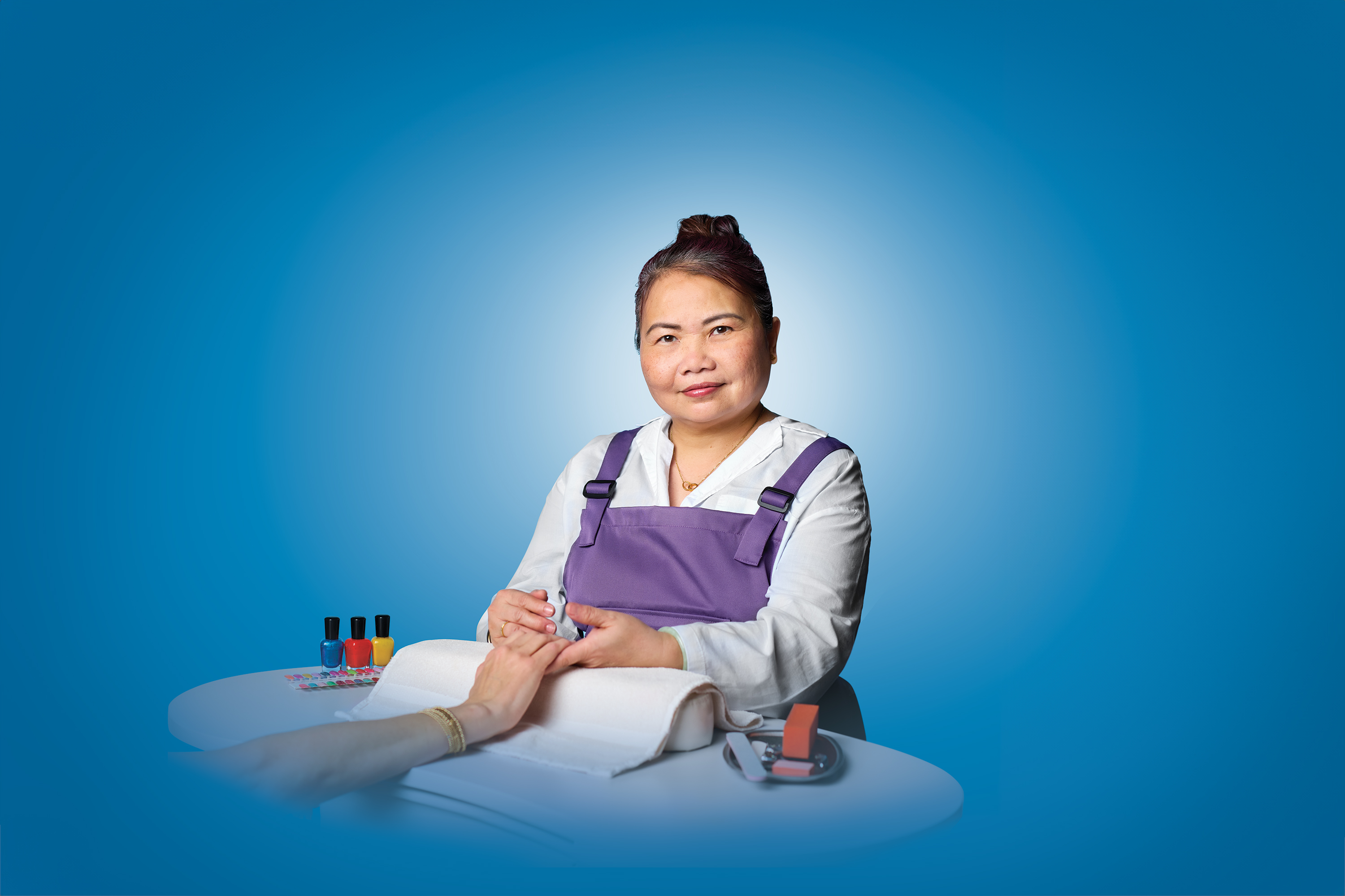 A sitting nail salon technician looks at the camera with a slight smile, wearing a purple apron 