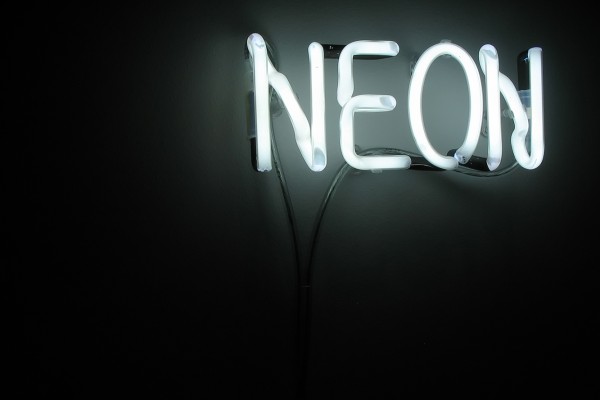 The image shows a neon sign with the word "NEON" lit up in white light. The sign is mounted on a dark wall and is the only source of light in the image, casting a soft glow around the letters. The neon tubes are arranged to spell out "NEON" in all capital letters, with the cabling and mounting structure faintly visible behind the glowing tubes