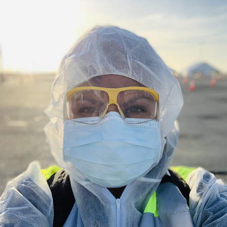 Woman wearing personal protective gear: white hair cap, blue mask, yellow-framed clear glasses, white suit 